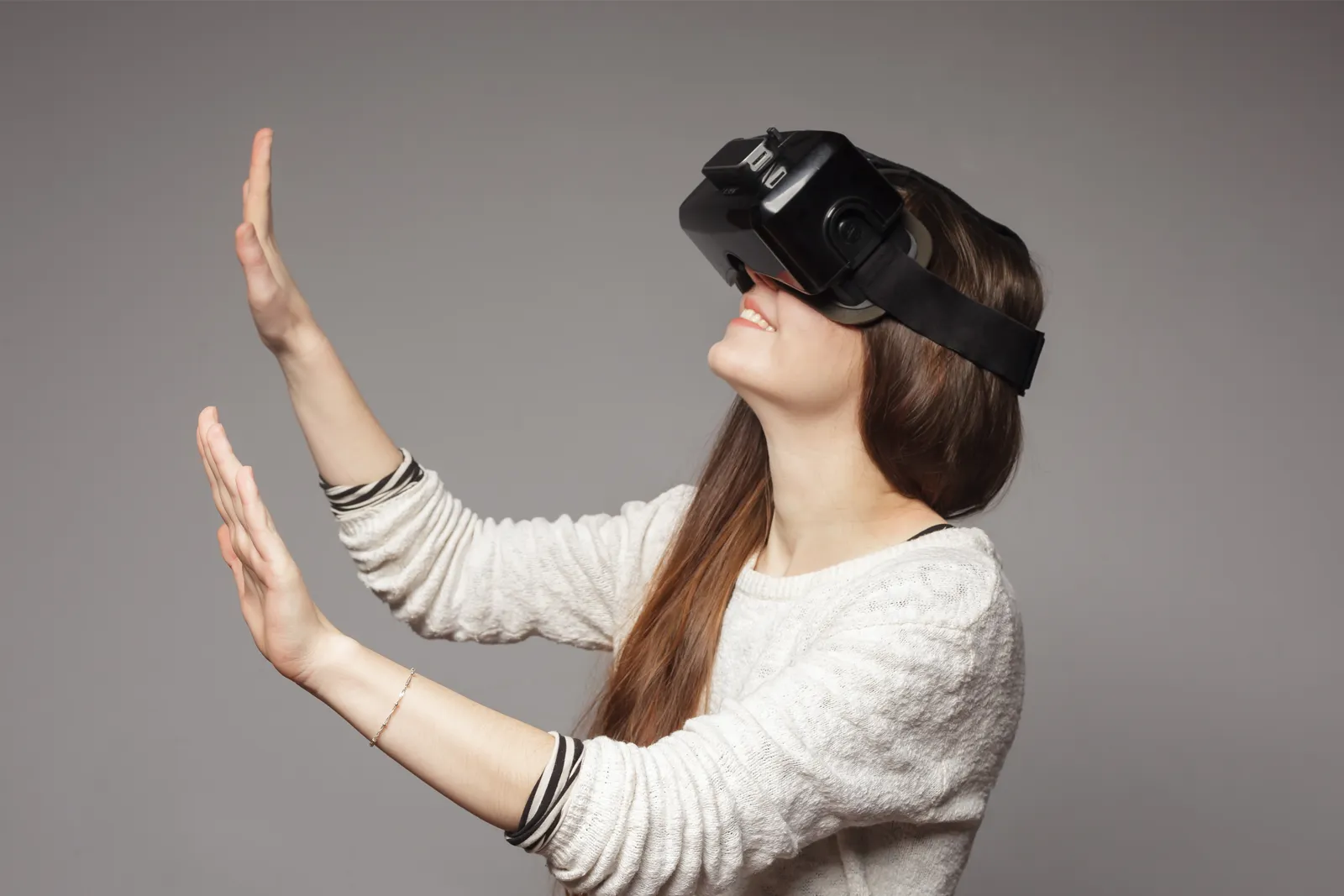 Mujer con gafas de realidad virtual negras y las manos levantadas sobre un fondo gris.